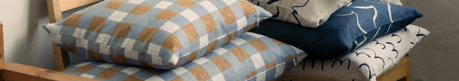 나무 의자에 쌓인 베개들, 체크무늬 파란색과 갈색, 그리고 추상적인 패턴의 파란색과 베이지색 디자인이 특징.
