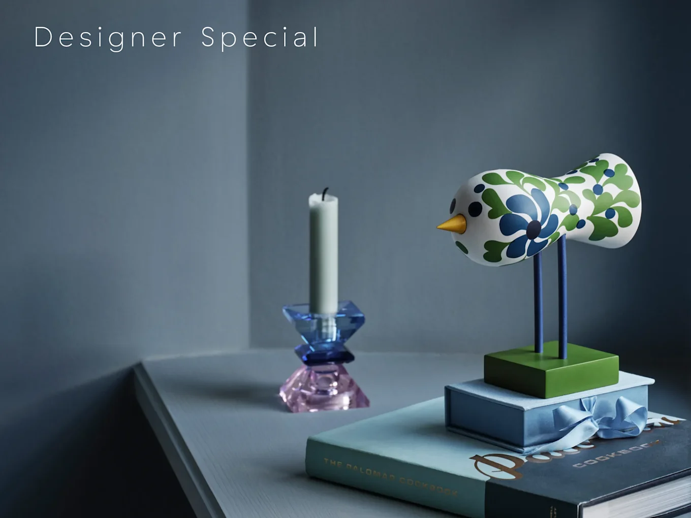 "Designer Special" 텍스트와 파란색, 녹색 무늬의 장식용 새 장식품, 두 가지 색상의 유리 촛대, 책들.