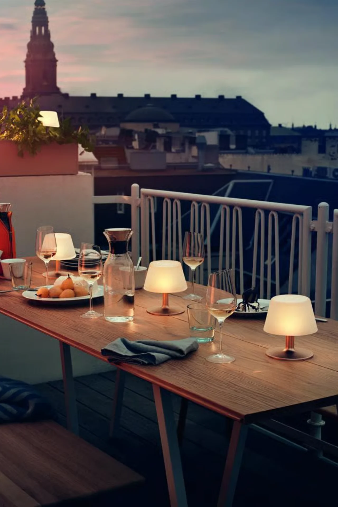 황혼녘 옥상 식탁에 빛나는 휴대용 램프, 와인, 과일, 물병이 놓여 있고 도시 스카이라인이 배경에 있다.