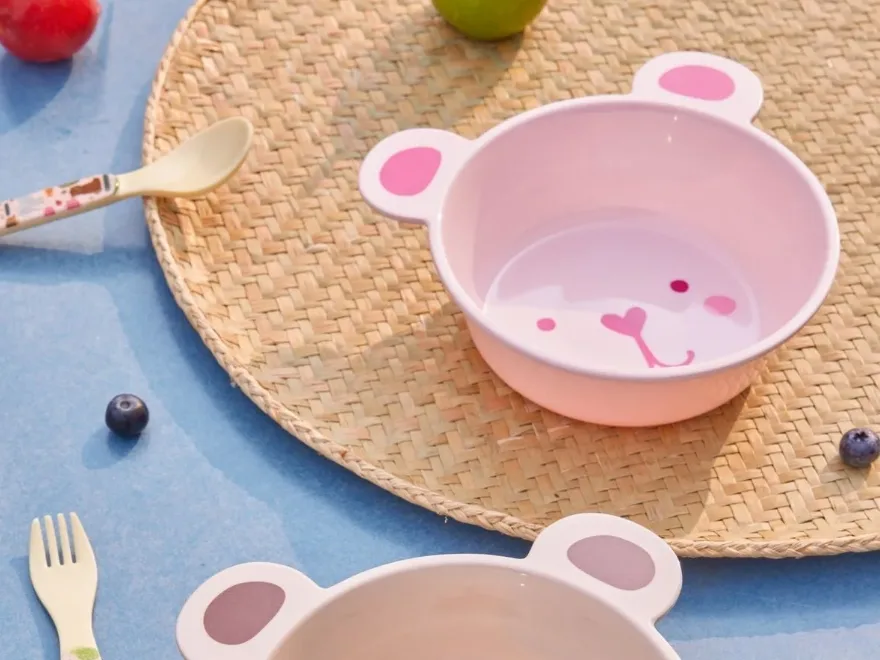 분홍색과 베이지색 곰 모양 그릇 두 개, 어울리는 숟가락과 포크, 과일과 함께 짜인 매트 위에 있습니다.
