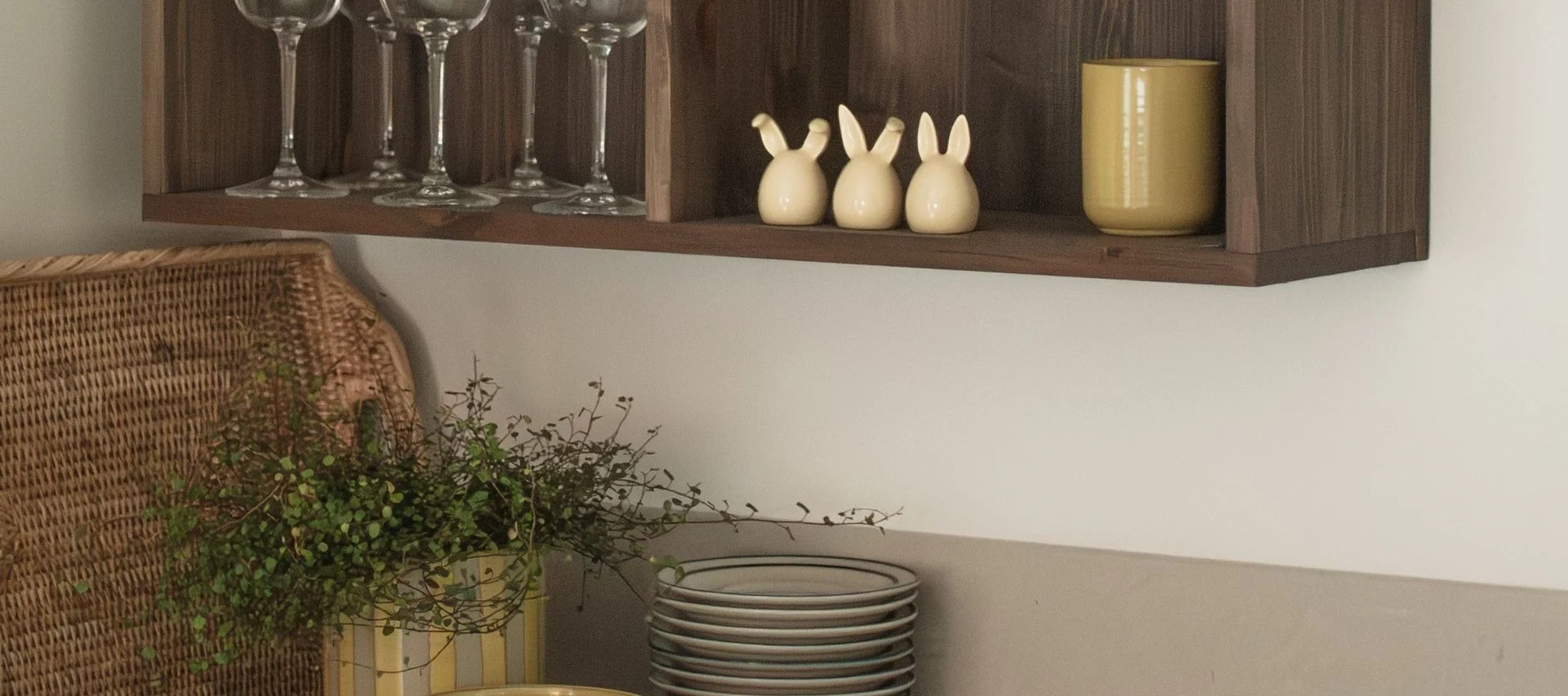 유리잔, 세 개의 토끼 장식품, 노란색 머그컵이 놓인 나무 선반과 그 아래에 있는 바구니와 식물.