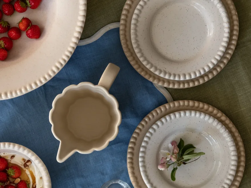 얼룩무늬 세라믹 접시, 딸기 그릇, 크리머가 파란색과 녹색 리넨 식탁보 위에 놓여 있고, 사과 꽃으로 장식되어 있습니다.