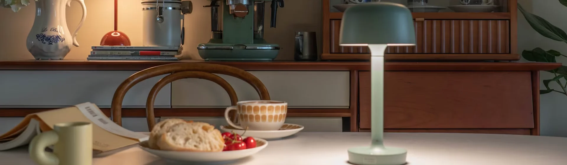 불이 켜진 연두색 램프, 커피, 빵, 체리가 있는 식탁이 아늑한 주방 배경에 놓여 있습니다.