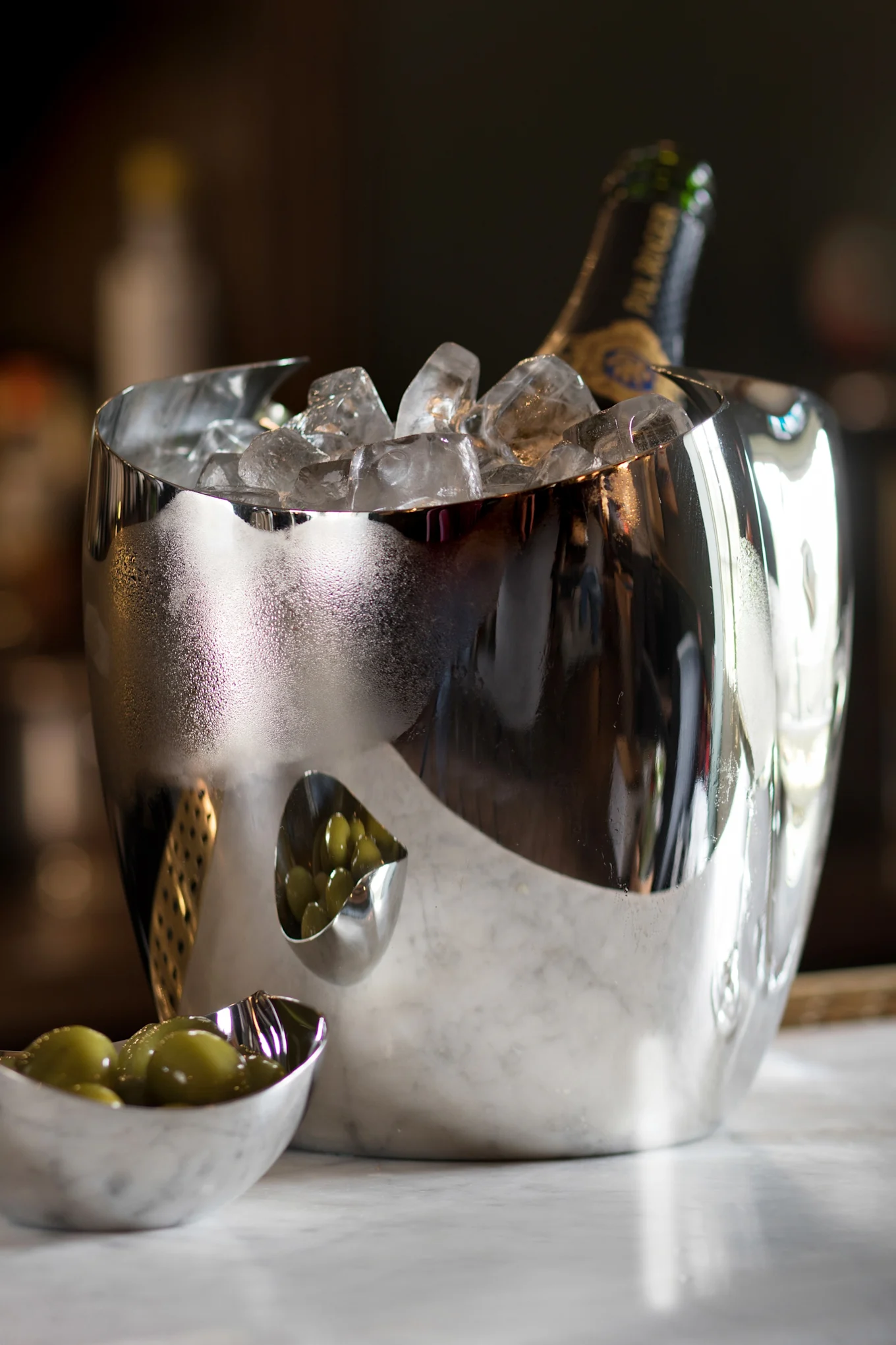 반사되는 은색 아이스 버킷에 샴페인 병과 얼음이 있고, 옆에는 대리석 위에 초록색 올리브가 담긴 작은 그릇 두 개가 있습니다.