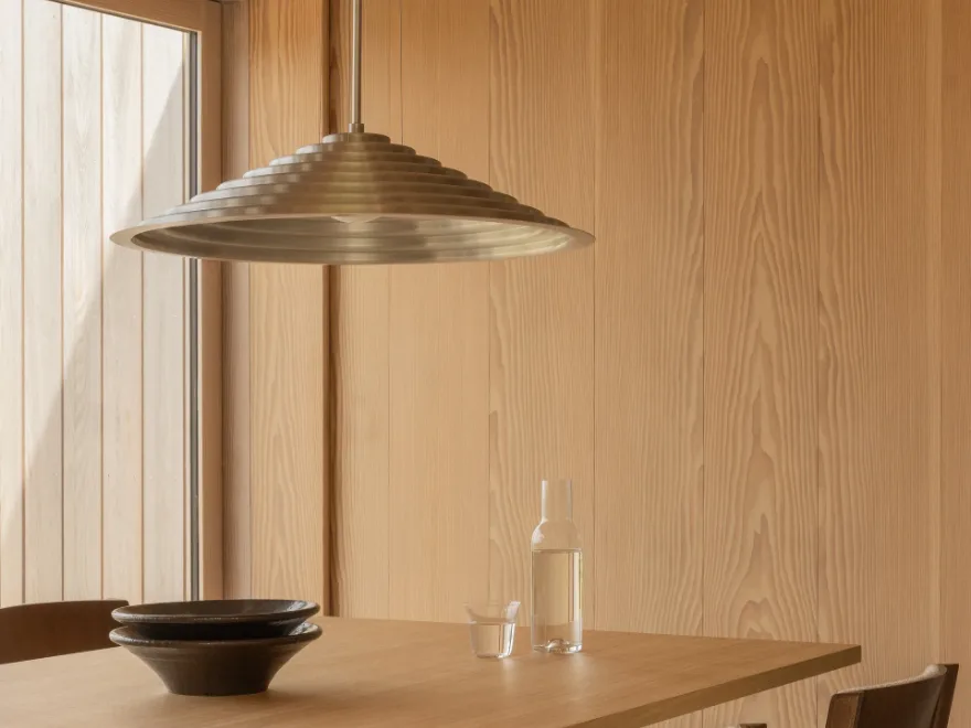 놋쇠색의 계단형 원뿔형 펜던트 조명이 밝은 나무 테이블 위에 걸려 있으며, 테이블 위에는 쌓인 어두운 세라믹 그릇, 물병, 유리잔이 있다.