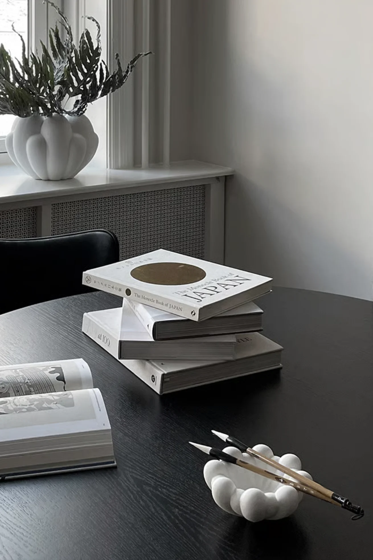어두운 테이블 위에 'The Monocle Book of Japan'을 포함한 흰색 책 더미와 구름 모양 그릇에 담긴 붓글씨 붓.