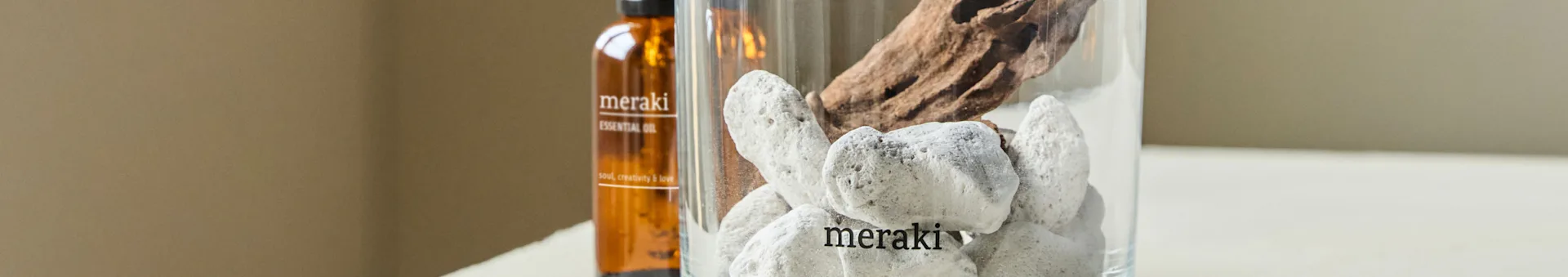 갈색 에센셜 오일 병과 흰 돌, 유목이 담긴 투명한 꽃병. 병과 꽃병 모두 'meraki'라고 라벨링되어 있습니다.